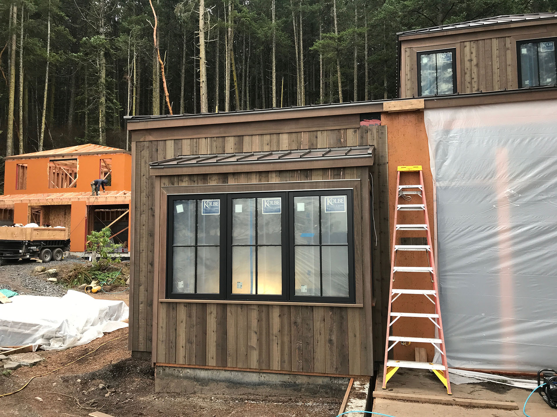 Construction site of a residential building with wooden framing and partially installed windows, surrounded by construction materials and equipment.