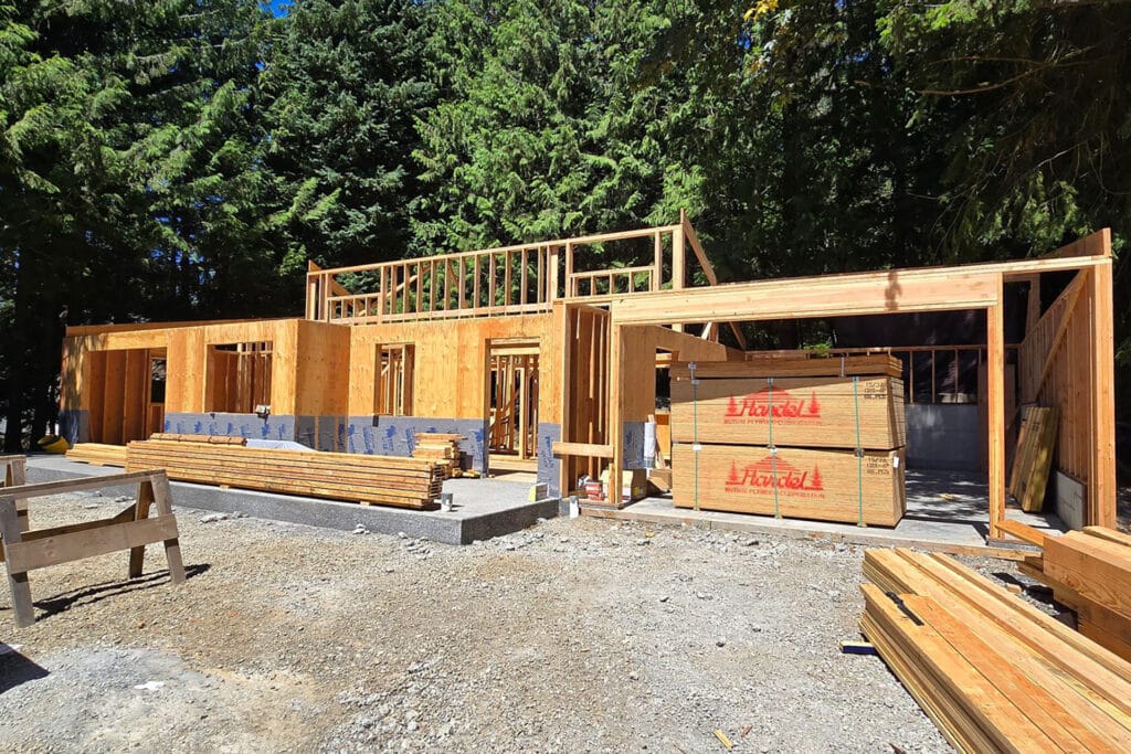 Exterior view of a residential construction site with wooden framing, showcasing the structure of walls and roof under a clear sky.